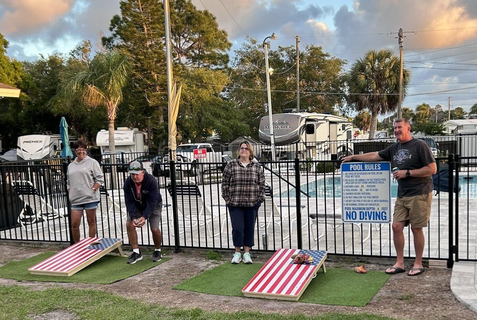 Children playing a bean bag toss game outdoors near parked boats.